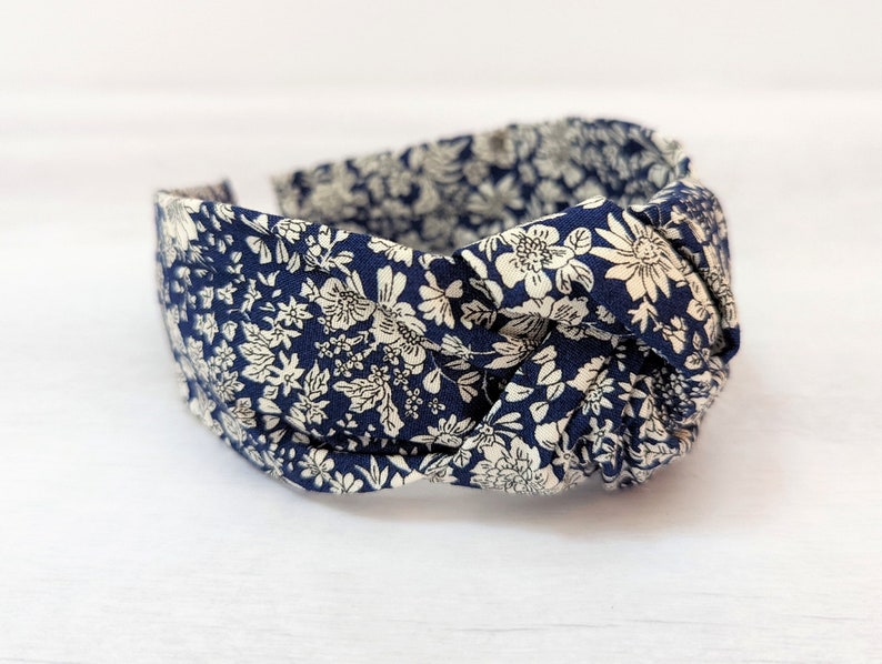 May include: A navy blue headband with a white floral pattern. The headband is knotted on top and is made of fabric.