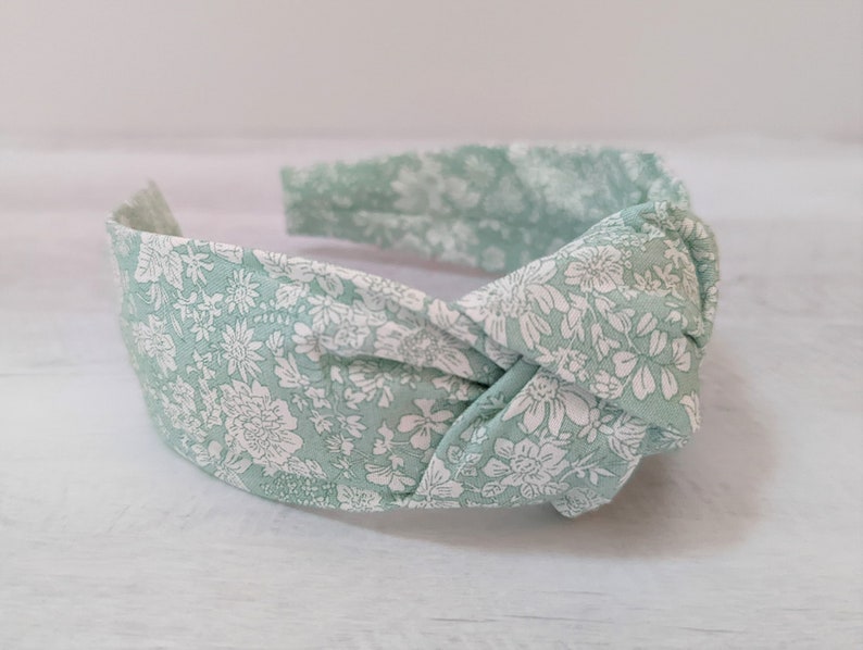 May include: A green headband with a white floral pattern. The headband is knotted in the front.