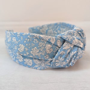 May include: A light blue headband with a white floral pattern. The headband is knotted on top and is made of fabric.