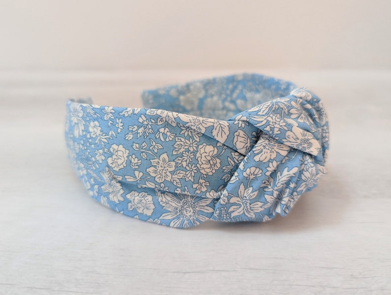 May include: A light blue headband with a white floral pattern. The headband is knotted on top and is made of fabric.