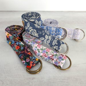 May include: Four fabric belts rolled into coils, each featuring a distinct floral pattern in shades of navy blue, dark purple, light pink, and lavender. Each belt is finished with a pair of metal D-rings at one end for secure fastening.