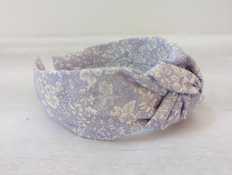 May include: A light purple fabric headband with a white floral pattern. The headband is knotted on top.