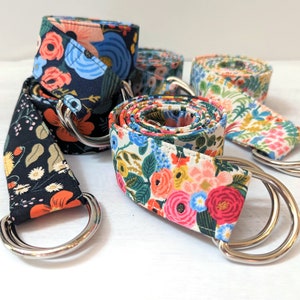 May include: Four fabric belts with floral patterns and silver D-ring buckles. The belts are rolled up and arranged on a white surface.