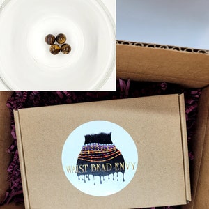 May include: A brown cardboard box with a circular sticker that reads "WAIST BEAD ENVY". The box contains purple shredded paper and a small glass bowl with four tiger eye beads. The box is inside a larger cardboard box.