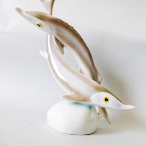 Könnte beinhalten: Eine Keramikskulptur von drei Delfinen. Die Delfine sind weiß und hellbraun mit gelben Augen und in einer dynamischen, aufwärts gerichteten Komposition angeordnet. Die Skulptur ist auf einem weißen Sockel montiert.