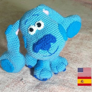 Puede incluir: Un juguete de peluche de perro azul tejido a crochet con ojos blancos, nariz azul y orejas rosas. El juguete está sentado sobre una superficie marrón claro.