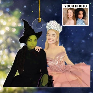 May include: A Christmas ornament featuring a green-skinned witch wearing a black hat and a pink-dressed princess wearing a tiara. The ornament is made of clear acrylic and has a gold string for hanging.