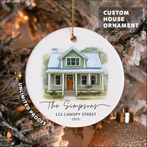 May include: A round white ceramic ornament featuring a watercolor illustration of a green house with a blue roof and a brown door. The text reads The Simpsons, 123 CANOPY STREET, and 2026. It hangs from a gold chain against a blurred tree background.