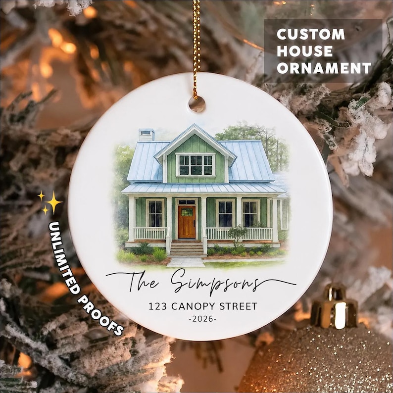 May include: A round white ceramic ornament featuring a watercolor illustration of a green house with a blue roof and a brown door. The text reads The Simpsons, 123 CANOPY STREET, and 2026. It hangs from a gold chain against a blurred tree background.