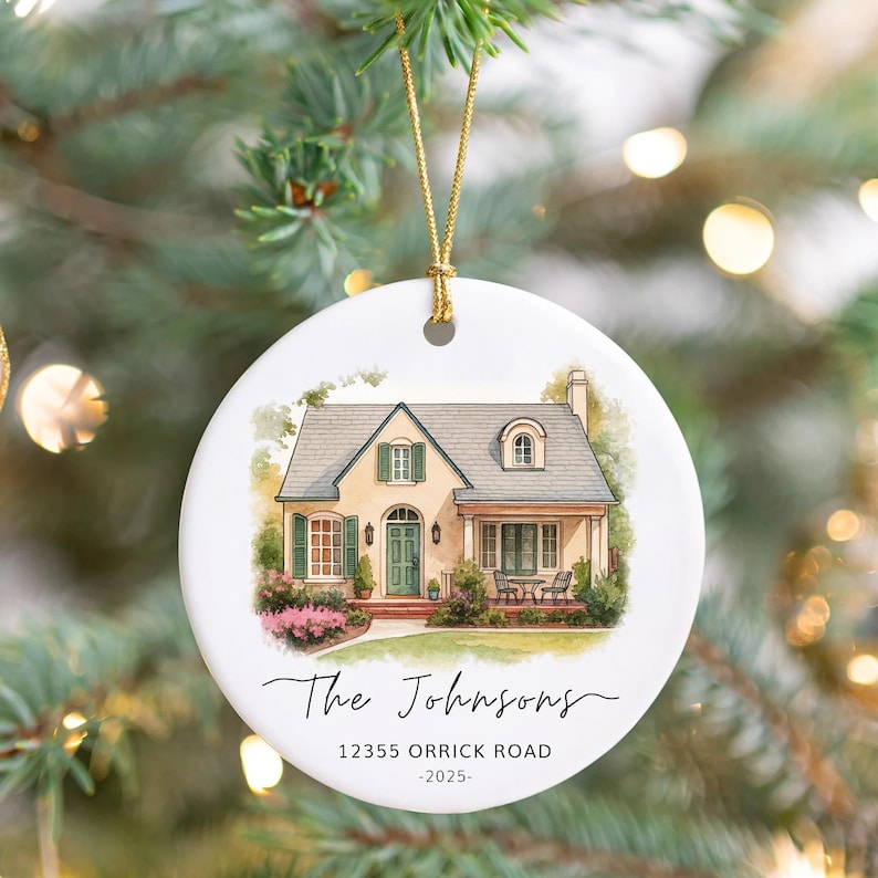 May include: A round white ceramic ornament featuring a watercolor illustration of a beige house with green shutters and a front porch. The text reads The Johnsons, 12355 ORRICK ROAD, and 2025. It hangs from a gold string against a blurred tree.