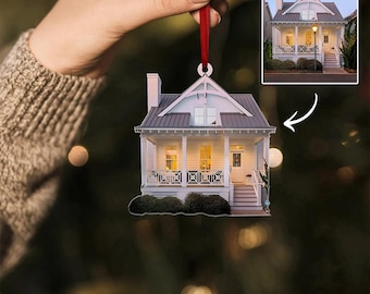 Pode incluir: Um ornamento de casa de estilo cottage branco com uma fita vermelha. O ornamento é um recorte detalhado de uma casa com uma varanda e janelas. A casa é iluminada com luzes amarelas quentes.