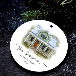 May include: A round white ceramic ornament featuring a watercolor illustration of a green house with a blue metal roof and a wooden front door. The text reads The Simpsons, 123 CANOPY STREET, and -2025- in black script and sans-serif fonts.