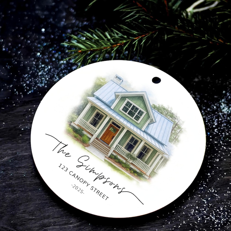 May include: A round white ceramic ornament featuring a watercolor illustration of a green house with a blue metal roof and a wooden front door. The text reads The Simpsons, 123 CANOPY STREET, and -2025- in black script and sans-serif fonts.