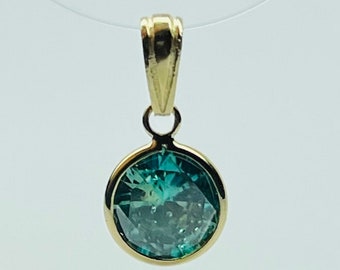 May include: A gold pendant with a round, faceted teal gemstone. The gemstone is set in a gold bezel and hangs from a gold bail. The pendant is suspended on a thin, clear cord.