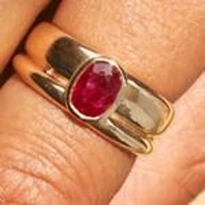 May include: A gold ring with a large, oval-shaped ruby gemstone. The ring has a thick band and a simple, elegant design. The ruby is a deep red color and is set in a gold bezel.