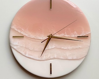 May include: A round wall clock with a two-tone design. The top half is a gradient of pink, while the bottom half is white. The clock features gold-colored hands and markers, creating a modern and elegant aesthetic. The clock's design resembles a beach scene.
