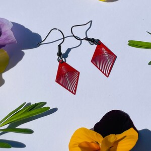 May include: A pair of red diamond-shaped earrings with a geometric design. The earrings have a black metal hook.