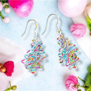 May include: A pair of silver dangle earrings with colourful, patterned, floral designs. The earrings are hanging against a light blue background.
