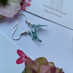 May include: A pair of silver hoop earrings with green fairy charms. The earrings are on a white background with the text "Enchanting Earrings".