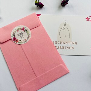 May include: A pink envelope with a floral thank you sticker. The envelope is on a white surface with a card that says "Enchanting Earrings" in brown text.