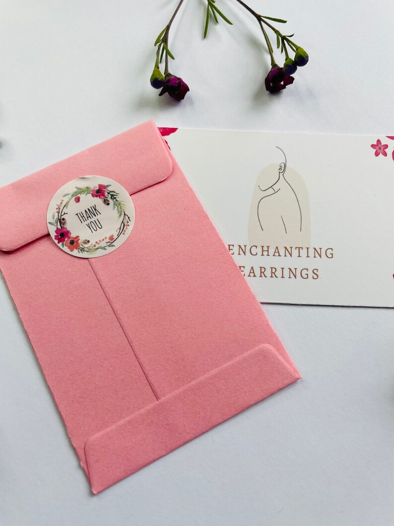 May include: A pink envelope with a floral thank you sticker. The envelope is on a white surface with a card that says "Enchanting Earrings" in brown text.