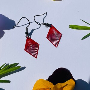 May include: A pair of red diamond-shaped earrings with a geometric design. The earrings have a black metal hook.