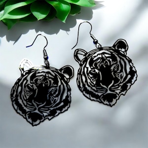 Può includere: Un paio di orecchini a testa di tigre in bianco e nero. Gli orecchini presentano un contorno dettagliato del volto di una tigre, con strisce e lineamenti del viso. Gli orecchini pendono da ganci per orecchie neri con una piccola perla blu.