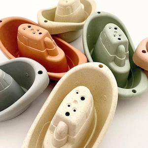 May include: Five small, colourful bath toys shaped like boats. The boats are made of a textured, rubber-like material and have holes in the top for water to drain. The boats are in various shades of green, beige, and orange.