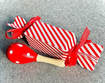May include: A red and white striped gift box tied with a red ribbon, resembling a large candy. Beside it is a red maraca with white polka dots and a natural wood handle. The items are on a gray surface.