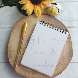 Op de afbeelding: Een wit notitieboekje met een gouden pen op een houten onderzetter. Het notitieboekje heeft de tekst "Gifts with love & smiles" erop geschreven. Een gehaakte bij zit op de onderzetter.