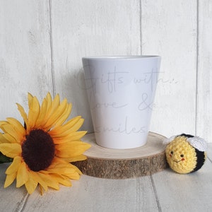 Puede incluir: Maceta de cerámica blanca con el texto "Gifts with love & smiles" en el lateral. La maceta está colocada sobre una rodaja de madera. En primer plano hay un girasol amarillo y una abeja de crochet.