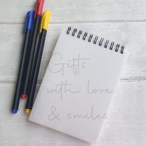 Puede incluir: Un bloc de notas blanco con encuadernación en espiral y el texto "Gifts with love & smiles" escrito en gris. Tres marcadores, uno rojo, uno azul y uno amarillo, están sobre una superficie de madera blanca.