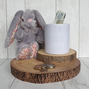 Könnte beinhalten: Eine weiße Keramik-Sparbüchse mit dem Text "Gifts with Love & Smiles" darauf. Die Spardose ist mit Bargeld gefüllt und steht auf einem Holzsockel, neben dem ein graues Plüschkaninchen sitzt.