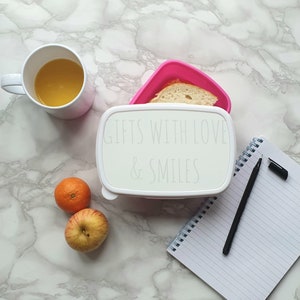 May include: White lunchbox with the text "Gifts with love & smiles" on the lid. The lunchbox is open and contains a slice of bread. There is a notebook, a pen, an orange, and an apple on a white marble surface.