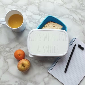 May include: White lunchbox with the text "Gifts with Love & Smiles" on a marble surface with a cup of orange juice, two mandarin oranges, an apple, a notebook, and a pen.