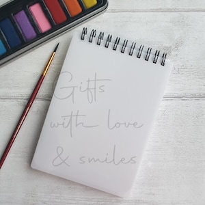 May include: A white spiral bound notebook with the words "Gifts with love & smiles" written in gray on the front. A paintbrush and a watercolor palette with colorful paints are on the table next to the notebook.