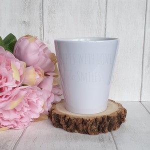 Könnte beinhalten: Ein weißer Keramik-Pflanztopf mit dem Text "Gifts with Love & Smiles" an der Seite. Der Topf steht auf einer Holzscheibe.