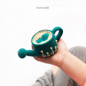 以下が含まれることがあります： ベージュのハートのデザインとベージュの花のアクセントが付いた、ターコイズブルーの編み物製のジョウロ。ジョウロは手で持たれています。