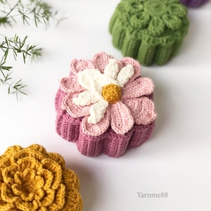 以下が含まれることがあります： 異なる色の花の形をした4つの編み物ボックス。紫、緑、白の花が付いたピンク、黄色。ボックスは白い表面の上に置かれています。右下隅にYarnme88と書かれています。