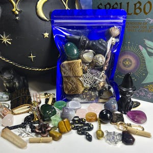 May include: A collection of crystals, charms, and small decorative items, including a black cat figurine, a blue bag of stones, and a spellbook. The items are arranged on a white surface, with a black cauldron in the background.