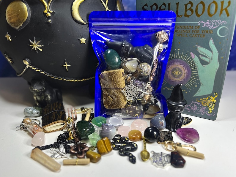 May include: A collection of crystals, charms, and small decorative items, including a black cat figurine, a blue bag of stones, and a spellbook. The items are arranged on a white surface, with a black cauldron in the background.
