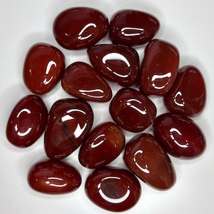 May include: A collection of polished, oval-shaped carnelian stones. The stones are a deep reddish-brown color with a glossy finish, reflecting light. The arrangement is scattered on a white surface, showcasing the natural variations in color and shape.
