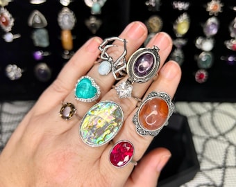 May include: A hand displays a collection of rings with various gemstones and designs. Rings include a heart-shaped turquoise stone, an oval abalone shell, and a ruby. Other rings feature clear, purple, and orange stones.