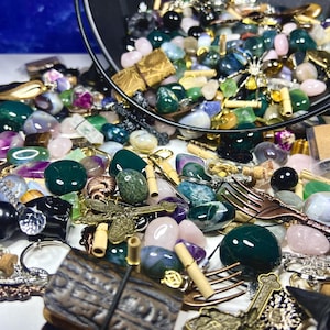 May include: A collection of gemstones, beads, and decorative items spilling from a black container. The assortment includes green, pink, and purple stones, along with metal charms and small forks. A variety of shapes and sizes are present.