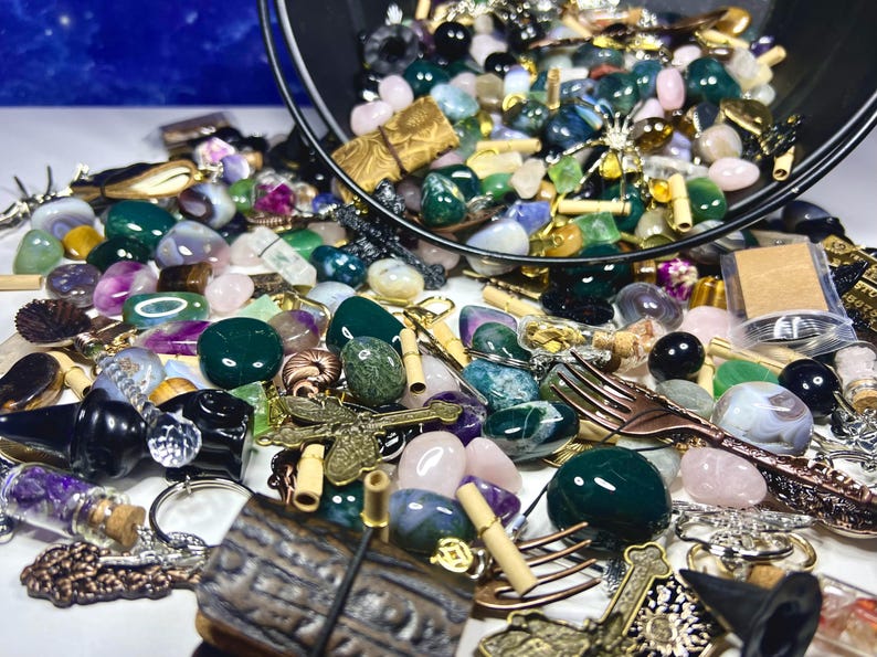 May include: A collection of gemstones, beads, and decorative items spilling from a black container. The assortment includes green, pink, and purple stones, along with metal charms and small forks. A variety of shapes and sizes are present.