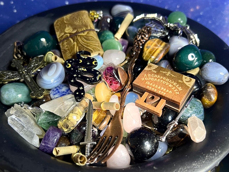 May include: A collection of various gemstones, charms, and small objects in a black bowl. Items include a black snake charm, a miniature Ouija board, a cross, a dagger, and a fork. The bowl is set against a starry background.