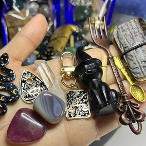 May include: A collection of mystical items including a black cat figurine wearing a witch hat, a snake pendant, a small journal, and various crystals. The items are held in a hand, showcasing their details and textures.