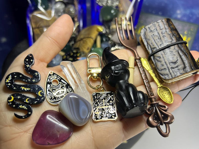 May include: A collection of mystical items including a black cat figurine wearing a witch hat, a snake pendant, a small journal, and various crystals. The items are held in a hand, showcasing their details and textures.