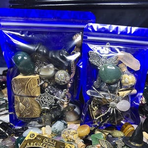 May include: Two blue zip-lock bags filled with assorted mystical items. The bags contain gemstones, charms, and small decorative objects. A variety of other items, including a Ouija board, are scattered around the bags.