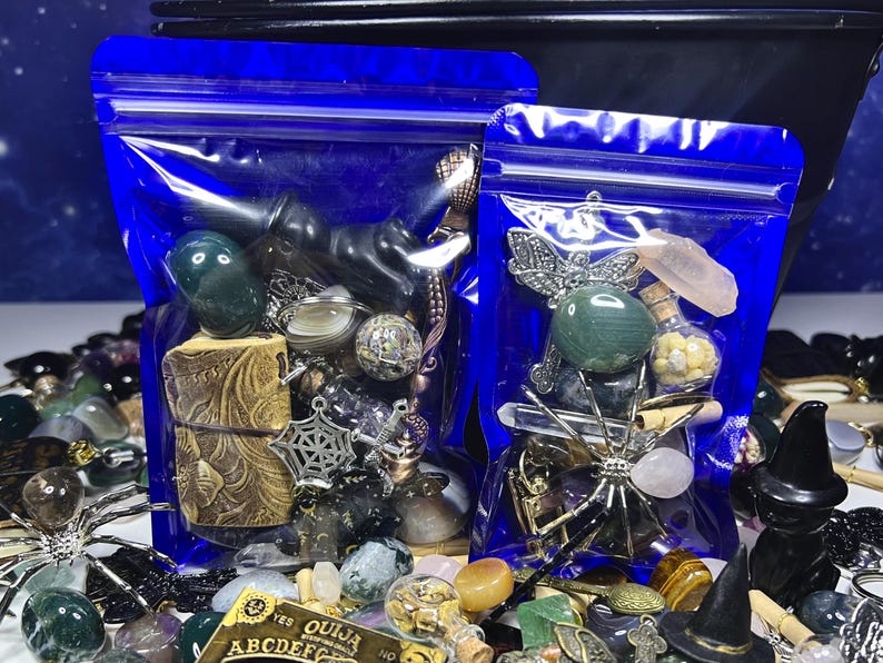May include: Two blue zip-lock bags filled with assorted mystical items. The bags contain gemstones, charms, and small decorative objects. A variety of other items, including a Ouija board, are scattered around the bags.